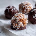 A close-up of delicious coconut balls rolled in shredded coconut, partially dipped in dark chocolate.