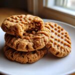 Close-up of stacked, classic fork-pressed peanut butter cookies, one with a bite taken out showing the soft interior.