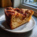 A close-up slice of moist jewish apple cake showing chunks of apple and a dark, caramelized topping.