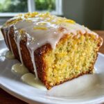 A close-up of a slice of moist lemon zucchini bread topped with thick white glaze and lemon zest.