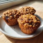 Three freshly baked, moist oatmeal muffins with visible oats and dried fruit sitting on a light speckled plate.