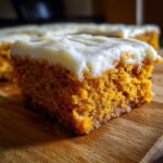 A moist, square slice of orange pumpkin bars with thick cream cheese frosting on a wooden board.