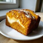 A thick slice of moist pumpkin cream cheese bread showing the orange pumpkin swirl and white cream cheese filling.