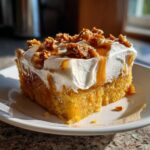 A moist slice of orange pumpkin poke cake topped with whipped cream, caramel drizzle, and candied pecans.