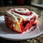 A close-up, appetizing shot of a single red velvet cinnamon roll topped with thick white cream cheese icing.