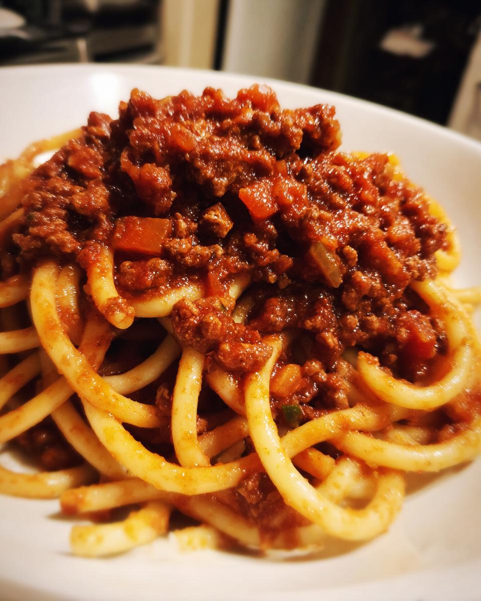 Close-up of thick, rich bolognese sauce generously spooned over a mound of bucatini pasta in a white bowl.