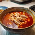Close-up of a rustic bowl filled with vibrant red chicken pozole, featuring shredded chicken and hominy kernels.