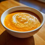 Close-up of vibrant orange roasted butternut squash soup, garnished with a swirl of cream and black pepper.