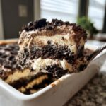 A spoonful of layered Oreo dirt cake showing chocolate, cream, and cookie crumb layers being lifted from a white dish.