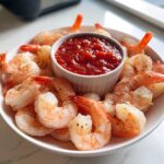 A white bowl filled with chilled, seasoned shrimp surrounding a central ramekin of red shrimp cocktail sauce.