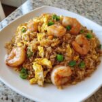 A close-up of a serving of shrimp fried rice recipe with large shrimp, scrambled egg, and green onions on a white plate.