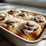 Close-up of warm, gooey cinnamon buns covered in thick white cream cheese frosting in a baking dish.