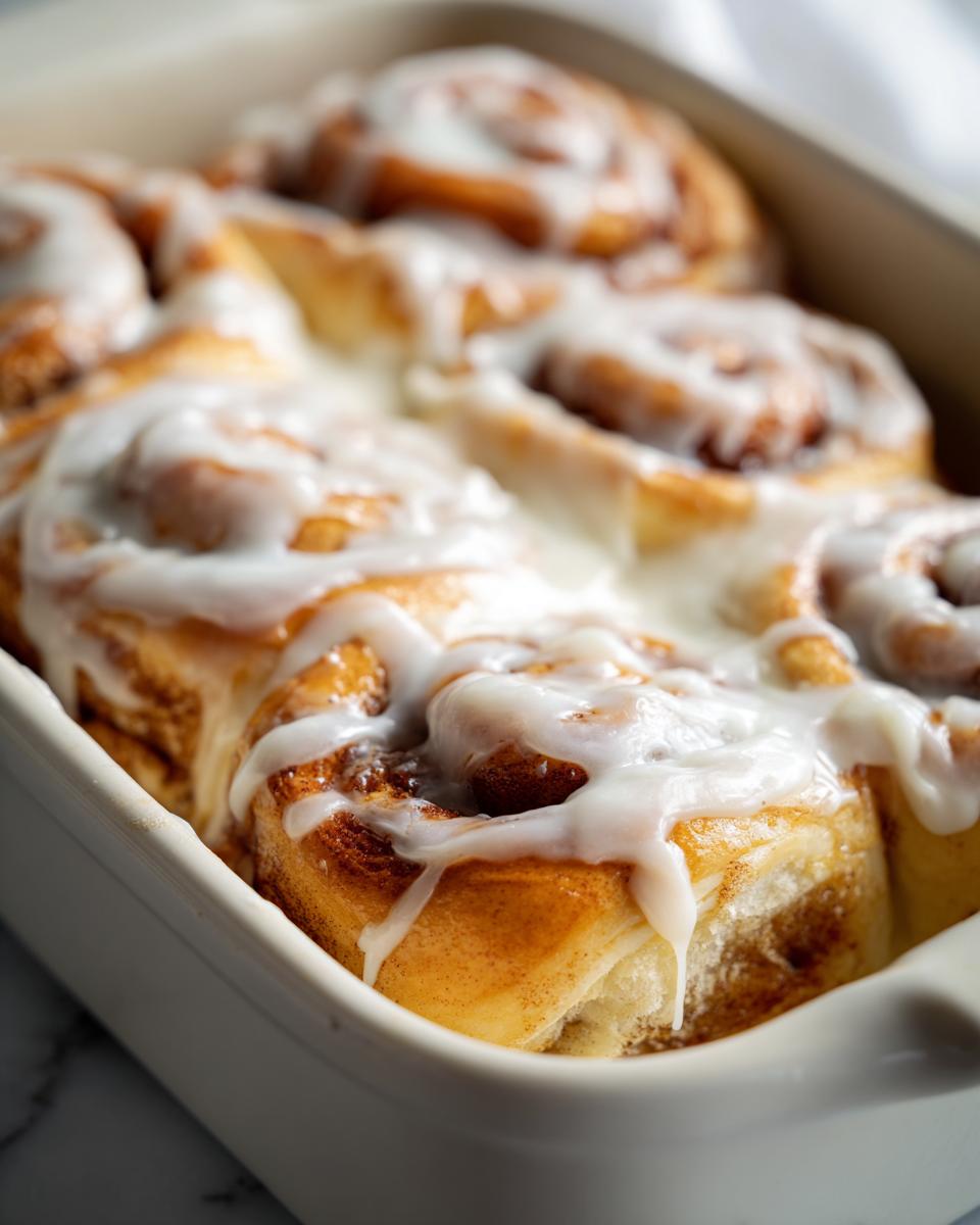 Close-up of soft, golden cinnamon buns baked together in a white dish, generously topped with dripping white icing.