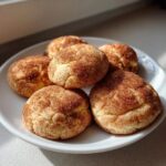 A close-up of five freshly baked snickerdoodle cookies piled on a white plate, coated in cinnamon sugar.