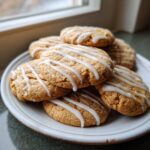 A stack of soft maple cookies drizzled with white icing, sitting on a light-colored plate near a window.