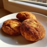 Three freshly baked, soft snickerdoodles heavily coated in cinnamon sugar resting on a white plate near a window.