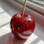 A close-up of a glossy, deep red candy apples coated in edible glitter, held by a wooden stick.