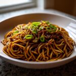 A close-up of a bowl filled with rich, dark sauce-coated spicy garlic noodles, topped with bright green scallions.