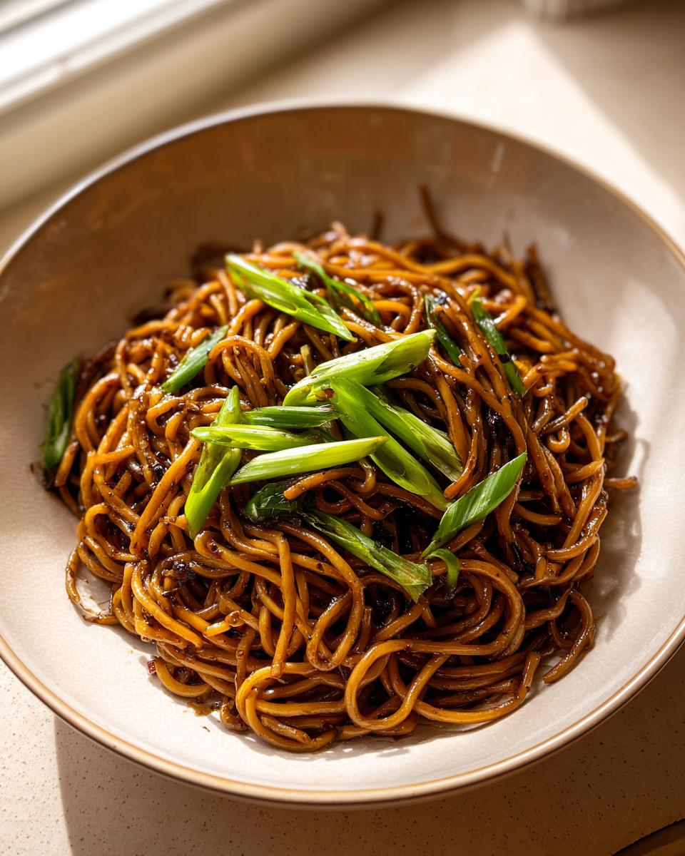 A close-up of glossy, dark brown spicy garlic noodles topped generously with bright green sliced scallions.