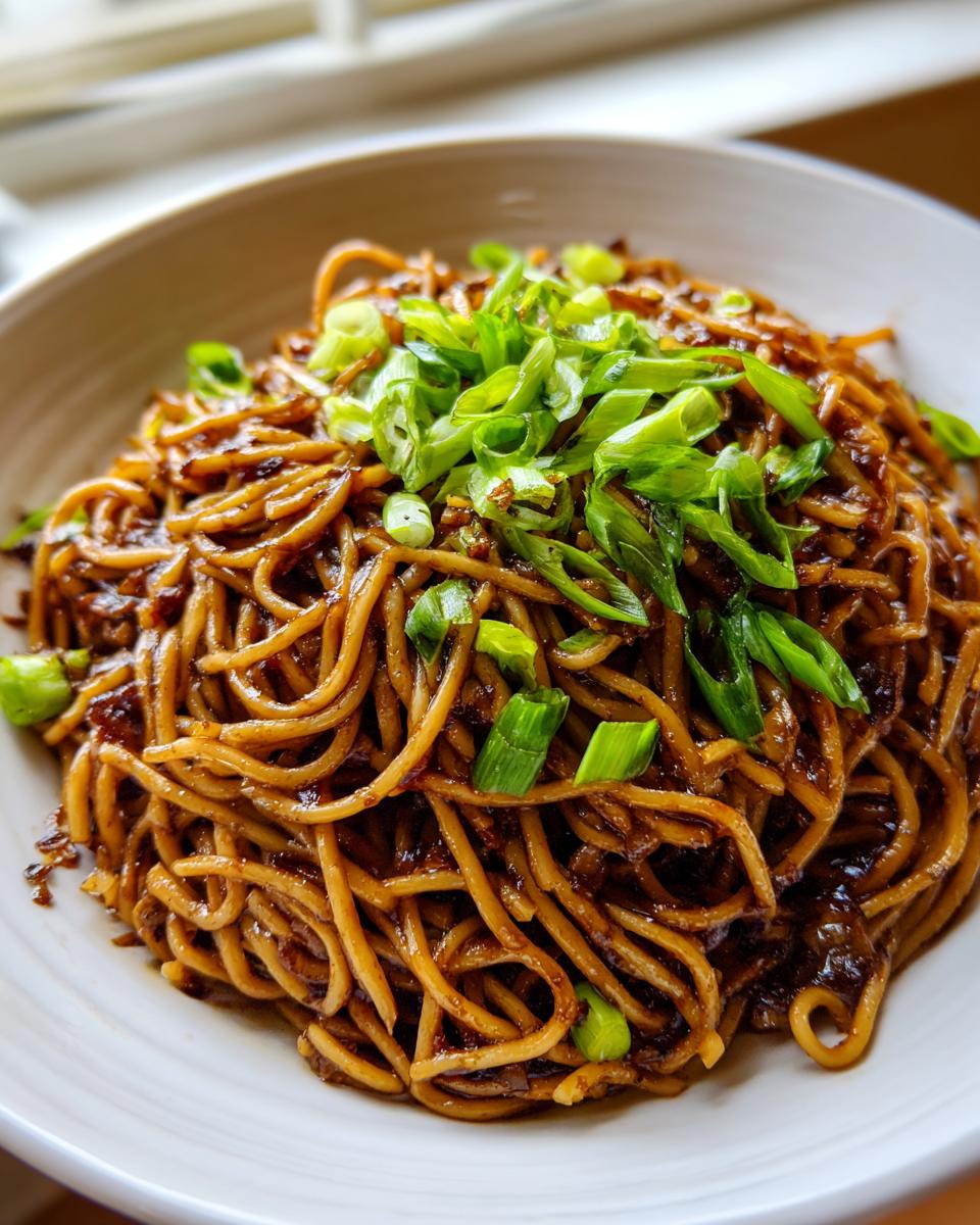 A close-up shot of a white bowl filled with saucy, dark brown spicy garlic noodles, topped generously with bright green sliced scallions.
