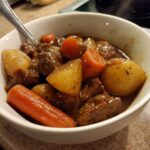 Close-up of a steaming white bowl filled with rich, dark pork stew chunks, potatoes, and bright orange carrots.