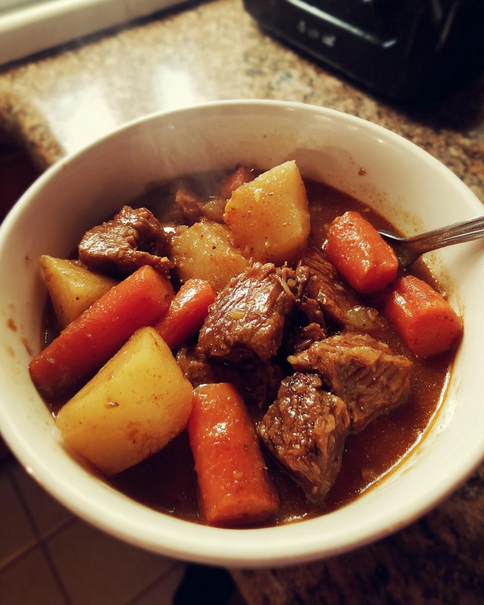 Close-up of a steaming white bowl filled with chunks of tender pork stew, potatoes, and carrots in rich gravy.