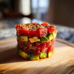A tall, cylindrical stack of vibrant red tuna tartare layered with green avocado cubes, topped with sesame seeds.