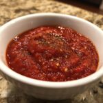 Close-up of thick, vibrant red pizza sauce seasoned with herbs and chili flakes in a small white bowl.