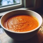 A close-up, cozy shot of a white bowl filled with vibrant orange tomato basil soup, set near a window.