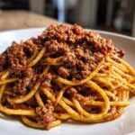 Close-up of spaghetti noodles generously topped with rich, dark bolognese sauce on a white plate.