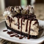 A decadent slice of ice cream cake with a dark cookie crust, creamy filling, chocolate syrup drizzle, and whipped cream topping.