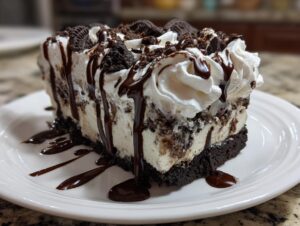 A decadent slice of 3-layer ice cream cake featuring cookies and cream filling, topped with whipped cream and chocolate syrup.