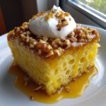 Close-up of a moist slice of butter pecan praline poke cake soaking in caramel sauce, topped with pecans and whipped cream.