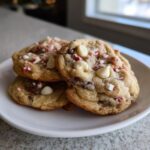 Three chewy peppermint chocolate chip cookies topped with white chocolate chips and crushed candy canes on a white plate.