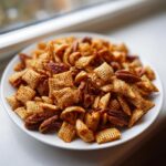 A close-up of seasoned chex party mix, including cereal squares and pecans, served on a white plate.
