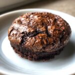 A single, rich, dark chocolate brownie cookie with a shiny, cracked top sitting on a white plate.