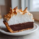 A decadent slice of chocolate meringue pie showing a thick, dark chocolate filling and a tall, fluffy, torched meringue topping.