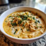 A close-up of a white bowl filled with rich, creamy cream cheese chicken chili, topped with shredded chicken, cheese, and fresh cilantro.