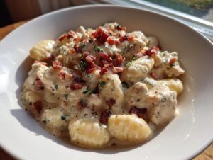 A close-up of creamy crack chicken gnocchi topped with crumbled bacon and herbs in a white bowl.