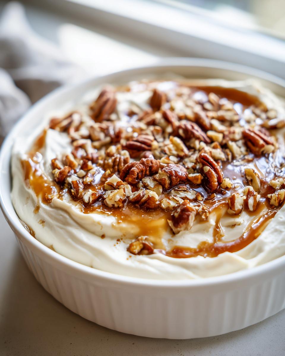 Close-up of a white bowl filled with creamy pecan pie dip, drizzled with caramel sauce and topped with whole and chopped pecans.