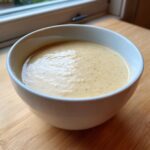 A close-up of a white bowl filled with thick, creamy roasted cauliflower soup, sitting on a wooden surface.