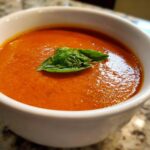 Close-up of a rich, orange-red bowl of homemade tomato bisque topped with a fresh basil leaf.