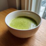 A close-up of a white bowl filled with vibrant green, creamy zucchini soup, sitting on a wooden table.