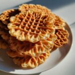 A close-up stack of freshly made, golden brown, crispy pizzelle cookies with a delicate waffle pattern, sitting on a white plate.