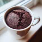 Close-up of a freshly baked, dark chocolate fudgy mug cake rising above the rim of a white ceramic mug.