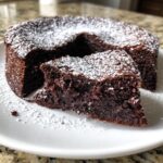 A rich, dark slice of flourless chocolate cake dusted heavily with powdered sugar, sitting next to the rest of the cake.