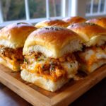 Close-up of several gooey buffalo chicken sliders with melted cheese and parsley on a wooden serving tray.