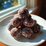 A pyramid stack of dark brown bourbon balls coated lightly in white powdered sugar on a white plate.