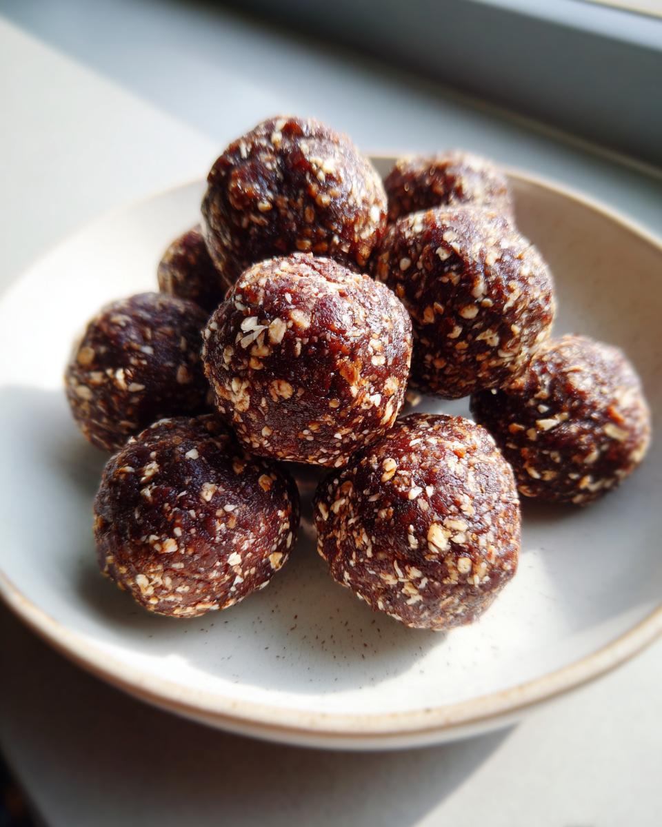 A close-up of several dark, textured protein balls sprinkled with visible oat flakes, stacked in a small bowl.