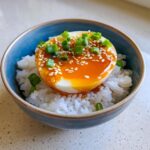 A halved, soft-boiled marinated egg served over white rice, topped with savory sauce, sesame seeds, and green onions.